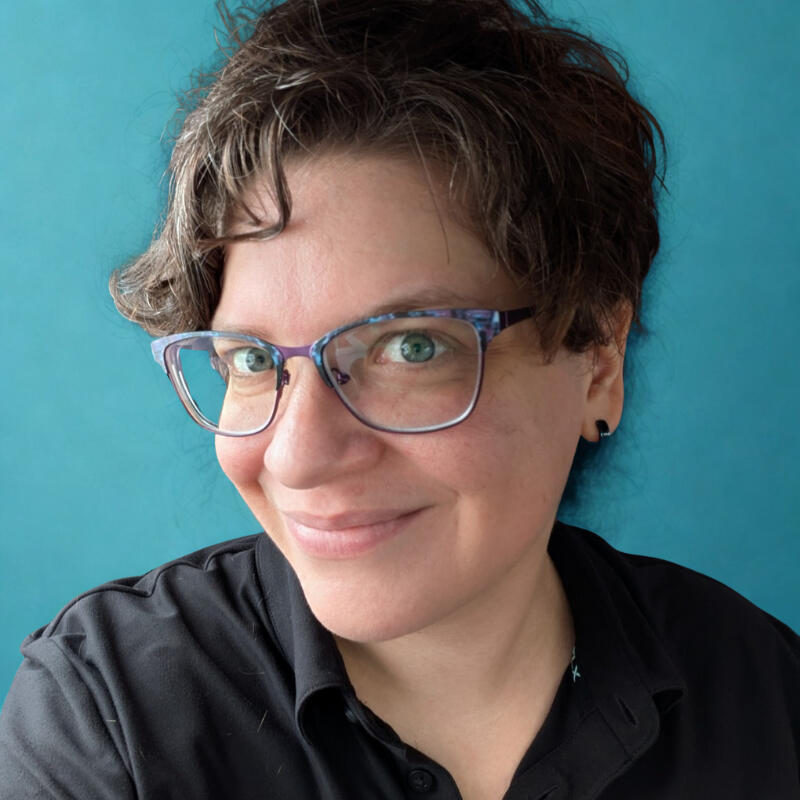 Natacha, Owner of Cosmic Spider (she/her/hers) A picture of Natacha, the owner of Cosmic Spider. She has short brown hair and blue eyes. She is smiling, wearing a black polo shirt, and purple and blue glasses.